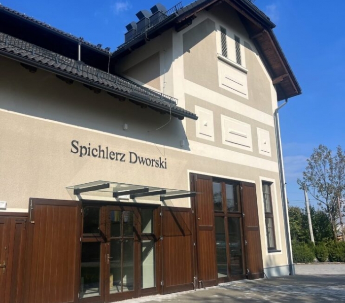 Budynek o jasnej elewacji z ciemnym dachem, dużymi drewnianymi drzwiami i przeszklonymi oknami na parterze. Przed budynkiem znajduje się kamienny podjazd, a w tle widać drzewa i niebo.